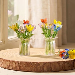 Pode incluir: Dois buquês de flores de vidro em vasos transparentes com bordas franzidas estão em uma fatia de madeira. As flores são coloridas, com pétalas amarelas, laranjas, roxas e azuis. Algumas flores soltas também estão na superfície de madeira.