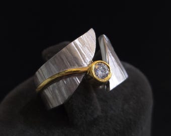 May include: A silver ring with a gold band and a bezel-set, round, clear gemstone. The ring has a unique design with textured, wing-like elements on either side of the gemstone, creating a modern and artistic look.