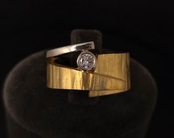 May include: A modern gold ring with a silver accent and a round, clear gemstone. The ring has a unique, geometric design with a textured gold band and a silver band that crosses over the top.