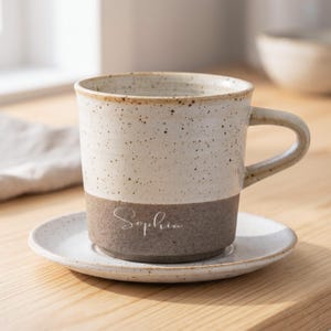 May include: A speckled ceramic mug with a matching saucer. The mug is two-toned, with a cream-colored upper half and a brown lower half. The name "Sophia" is written on the brown section. The mug has a handle and is sitting on a wooden surface.