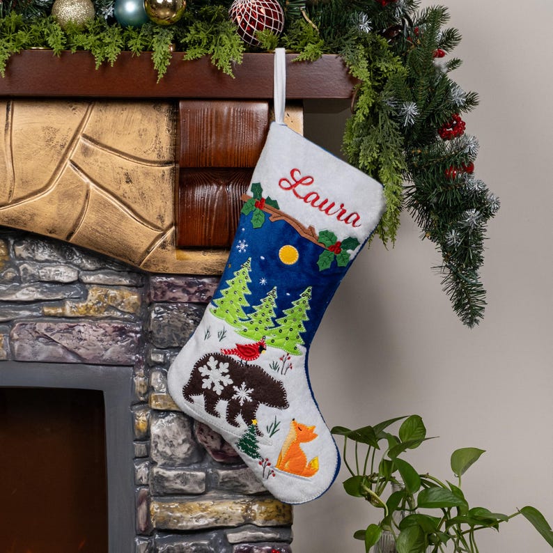 May include: A white and blue Christmas stocking featuring the embroidered name Laura in red script. The design includes a brown bear with white snowflake patterns, a bright orange fox, green pine trees, a yellow moon, and a red cardinal bird.