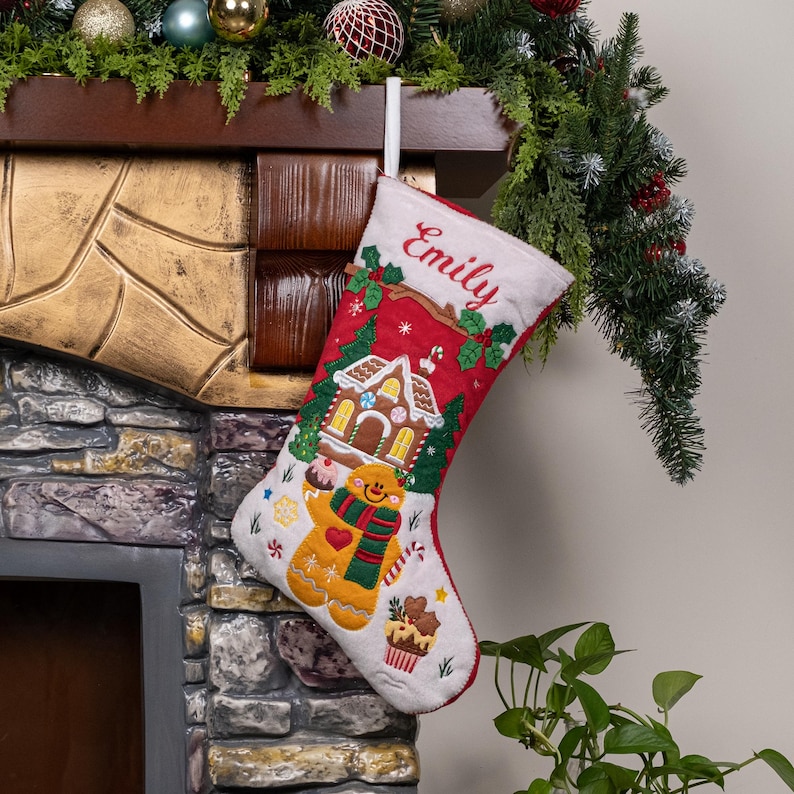May include: A white and red Christmas stocking featuring the embroidered name Emily at the top. The stocking displays a gingerbread house, a gingerbread person wearing a green and red scarf, and a cupcake, all decorated with festive holiday motifs.