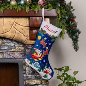 May include: A deep blue Christmas stocking with a white cuff featuring the embroidered name Hazel in red script. The stocking displays a colorful scene of Santa in a sleigh pulled by reindeer, a yellow moon, white snowflakes, and small houses.
