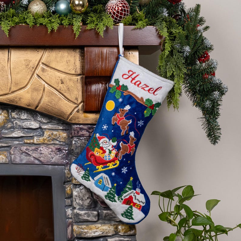 May include: A deep blue Christmas stocking with a white cuff featuring the embroidered name Hazel in red script. The stocking displays a colorful scene of Santa in a sleigh pulled by reindeer, a yellow moon, white snowflakes, and small houses.