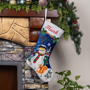 May include: A blue and white Christmas stocking featuring an embroidered snowman wearing a top hat and a red and green scarf, a squirrel, holly leaves, and pinecones. The name Hank is embroidered in red script at the top of the stocking.