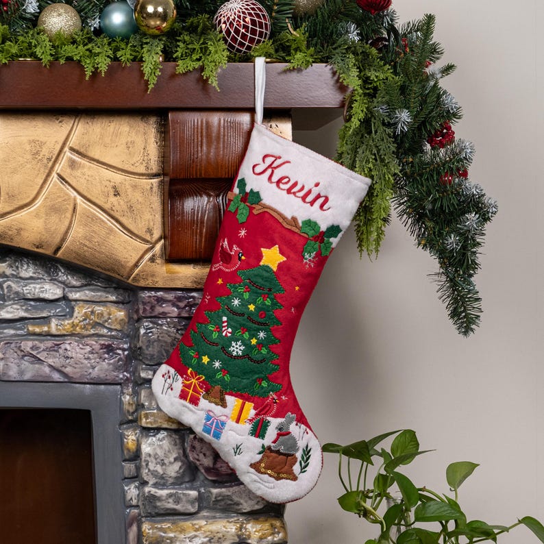 May include: A red Christmas stocking featuring the embroidered name Kevin at the top. The stocking displays a green Christmas tree, colorful gift boxes, a small gray rabbit, and holly leaves. It is made of fabric and includes a white hanging loop.