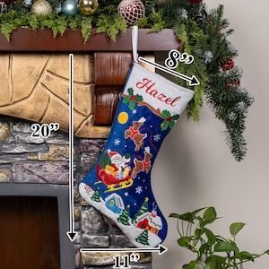 May include: A blue Christmas stocking with a white cuff embroidered with the name Hazel. The stocking features a festive scene with Santa in a sleigh, reindeer, a yellow moon, and snowy houses. It measures 20 inches in height, 8 inches at the top, and 11 inches at the base.