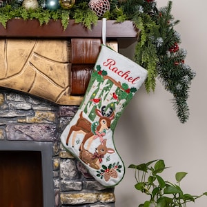 May include: A white Christmas stocking featuring an embroidered scene of two deer in a birch forest. The name Rachel is stitched in red at the top. The design includes green holly leaves, red berries, and small red birds on the tree branches.