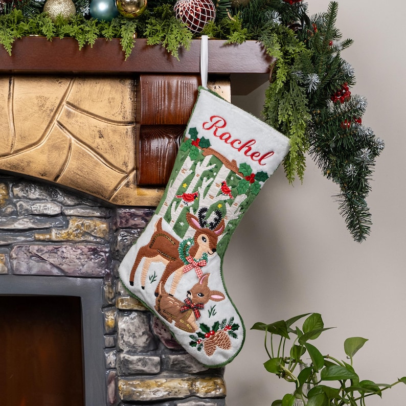 May include: A white Christmas stocking featuring an embroidered scene of two deer in a birch forest. The name Rachel is stitched in red at the top. The design includes green holly leaves, red berries, and small red birds on the tree branches.