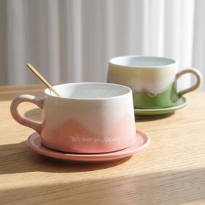 May include: Two ceramic mugs with saucers on a wooden surface. One mug is pink and white with the text "We love you, Mommy!" and a gold spoon. The other mug is green and white.