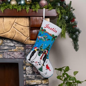 May include: A white Christmas stocking featuring the embroidered name Justin in red script at the top. The stocking displays a blue winter scene with two penguins wearing red Santa hats and scarves, green pine trees, and a red cardinal bird.