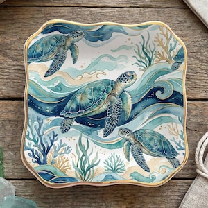 May include: A square ceramic trinket dish with scalloped edges and a gold-painted rim. The surface features a watercolor illustration of three sea turtles swimming among blue ocean waves, swirling currents, and various coral reef formations.