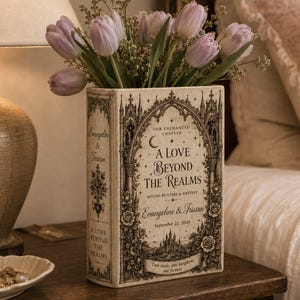 May include: A rectangular ceramic vase shaped like a book, featuring black gothic-style illustrations, floral motifs, and the text "A Love Beyond The Realms," "Evangeline & Tristan," and "September 21, 2026," filled with light purple tulips.