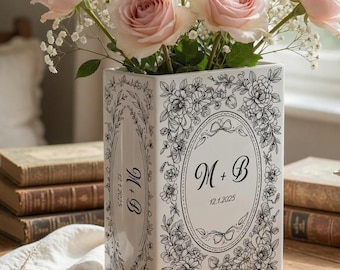 May include: A rectangular white ceramic vase with black line art floral patterns and an oval frame containing the text M + B and 12.1.2025. The side features the text M + B and 12.1.2025. The vase is filled with light pink roses and baby's breath.