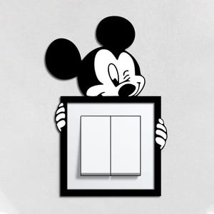 May include: A black and white Mickey Mouse decal holding a black framed light switch with two white switches. The cartoon character is winking and smiling, adding a playful touch to the wall decor.