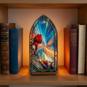 May include: A stained glass-style lamp featuring a figure in a red spacesuit climbing a structure, set against a vibrant blue, orange, and yellow background. The lamp is housed in a wooden frame and sits on a wooden base, resembling a church window.