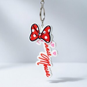 May include: A Minnie Mouse keyring featuring a red bow with white polka dots and the name "Minnie Mouse" in red script. The keyring includes a silver metal ring for attaching to keys or bags.