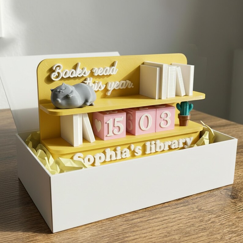 May include: A yellow and white bookshelf-shaped decorative item inside a white box. The shelf has the text "Books read this year" and "Sophia's library". It features a gray hippo figurine, white book stacks, and pink number blocks.