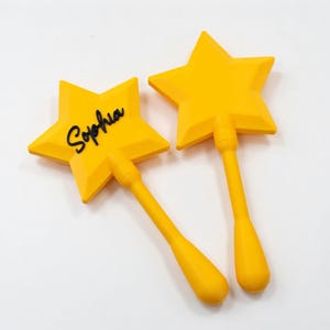 May include: Two yellow star-shaped wands with long handles. One wand has the name "Sophia" in black script. The wands are made of a solid material and are set against a white background. The wands are likely toys or decorative items.