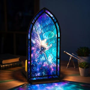 May include: A stained glass lamp in a gothic arch shape, featuring a dragonfly design against a backdrop of a nebula in shades of blue, purple, and teal. The lamp is illuminated, casting colorful light patterns. The lamp is on a wooden surface.