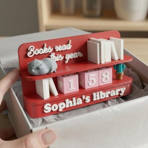 May include: A red bookshelf-shaped desk organizer with the text "Books read this year" and "Sophia's library". It features miniature books, a gray animal figurine, and pink number blocks. A small cactus is also present.