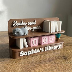 May include: A brown 3D-printed bookshelf with the text "Books read this year" and "Sophia's library". It features a gray hippo figurine, white book stacks, pink number blocks, and a small cactus. The blocks display the numbers 0603.