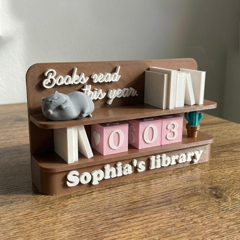 May include: A brown 3D-printed bookshelf with the text "Books read this year" and "Sophia's library". It features a gray hippo figurine, white book stacks, pink number blocks, and a small cactus. The blocks display the numbers 0603.