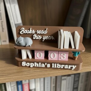 May include: A brown bookshelf-style organizer with the text "Books read this year" and "Sophia's library." It features a gray hippo figurine, white book-shaped dividers, and pink number blocks reading "003."