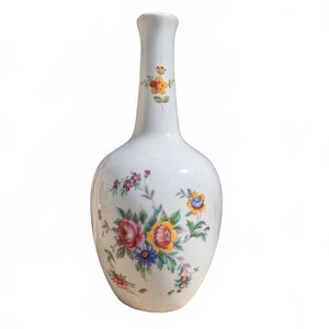 May include: A white ceramic vase with a narrow neck and a rounded body, featuring a colorful floral pattern with red, yellow, and blue flowers and green leaves painted on the front. The design includes a small floral sprig on the neck.