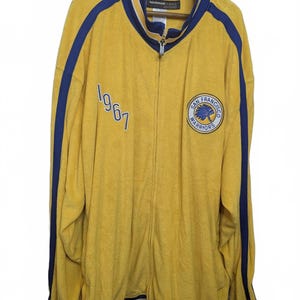 May include: A yellow zip-up track jacket with blue stripes along the sleeves and collar. The front features the embroidered text 1967 and a circular San Francisco Warriors logo patch. The garment has a high collar and ribbed cuffs and hem.