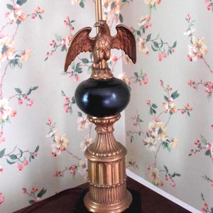 May include: A decorative table lamp with a bronze eagle perched atop a black orb. The lamp has a gold-colored base and a black square base. The lamp is approximately 30 inches tall.