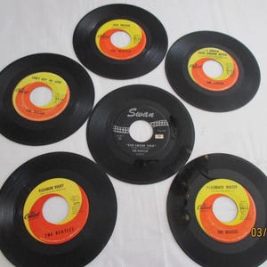 Puede incluir: Una colección de discos de vinilo vintage de The Beatles. Los discos son negros con etiquetas amarillas y naranjas. Los títulos incluyen "Can't Buy Me Love", "Eleanor Rigby" y "She Loves You."