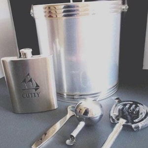 Può includere: Un set da bar in acciaio inossidabile con un secchiello per il ghiaccio, una fiaschetta con il logo "CUTTY" e strumenti da bar. Il secchiello per il ghiaccio ha un manico e un bordo decorativo. La fiaschetta è rettangolare. Gli strumenti da bar includono un jigger e uno strainer.
