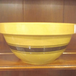 May include: A yellow ceramic mixing bowl with a wide rim and two dark brown stripes. The bowl is set against a wooden background, suggesting a kitchen or dining setting. The bowl's design is simple and functional.