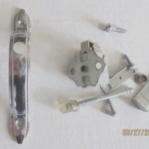 May include: A silver-toned metal door handle plate with two screw holes, accompanied by a collection of hardware components including a central latch mechanism, several small metal screws, a metal rod with a plastic tip, and mounting brackets.