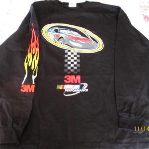 May include: Black long-sleeved shirt with a race car graphic in an oval, a chequered flag design, and the text "3M NASCAR PERFORMANCE." The left sleeve has a flame design and the "3M" logo. A classic style shirt.