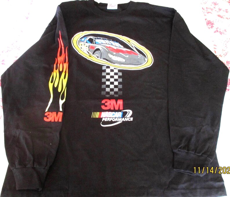 May include: Black long-sleeved shirt with a race car graphic in an oval, a chequered flag design, and the text "3M NASCAR PERFORMANCE." The left sleeve has a flame design and the "3M" logo. A classic style shirt.