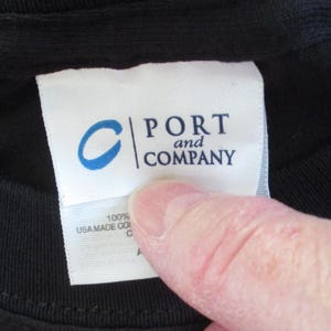 May include: A close-up of a black t-shirt with a white label. The label displays the blue "C" logo and the words "PORT and COMPANY". Below the logo, the label reads "100% USA MADE CO".
