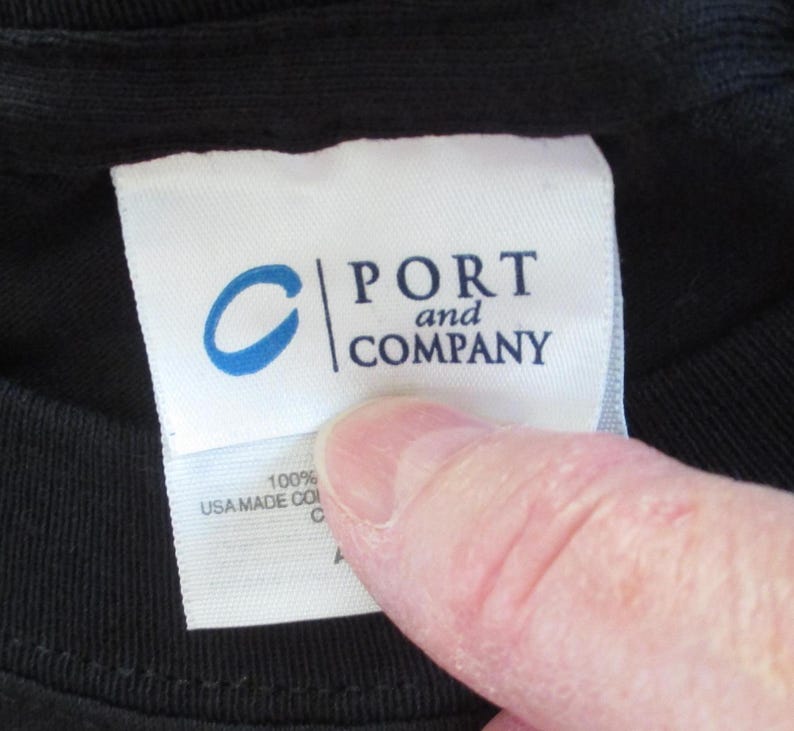 May include: A close-up of a black t-shirt with a white label. The label displays the blue "C" logo and the words "PORT and COMPANY". Below the logo, the label reads "100% USA MADE CO".