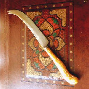 May include: A curved, silver-bladed knife with a light brown handle. The knife is set against a detailed, ornate background with red, gold, and green patterns. The handle has a textured appearance.