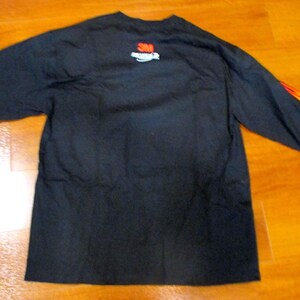 May include: Black long-sleeved shirt featuring a flame design on the right sleeve. The 3M logo is on the upper back. The date 12/08/2025 is visible. The shirt is displayed on a wooden surface.