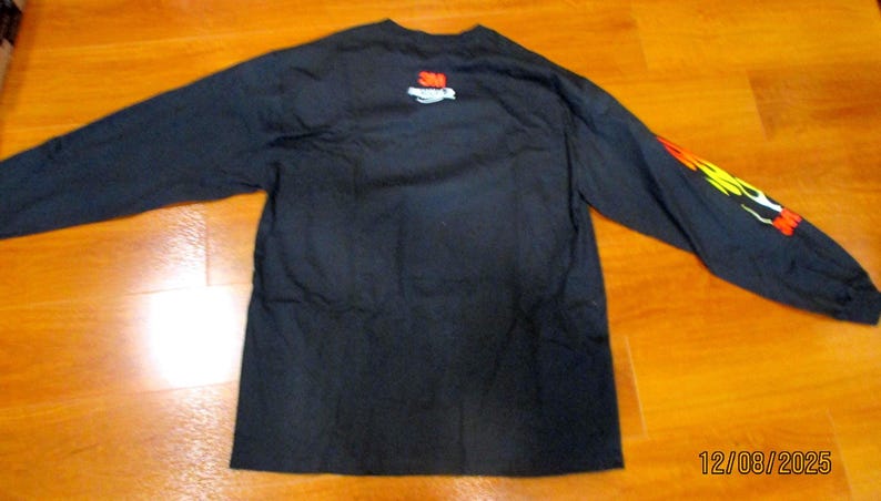 May include: Black long-sleeved shirt featuring a flame design on the right sleeve. The 3M logo is on the upper back. The date 12/08/2025 is visible. The shirt is displayed on a wooden surface.