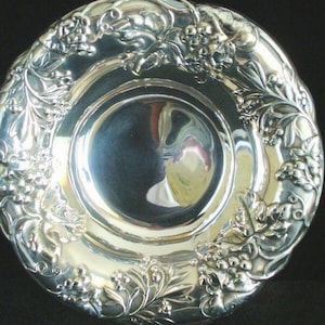 May include: A silver-colored plate with a detailed floral and grape vine design around the rim. The plate has a reflective surface and a slightly scalloped edge. The design is raised, creating a textured appearance. The plate is likely made of metal.