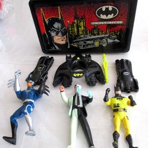 May include: A black Batman lunchbox with a handle, featuring Batman graphics and the text "Batman Returns." Includes Batman action figures and accessories, including a Two-Face figure and a yellow Batman figure.