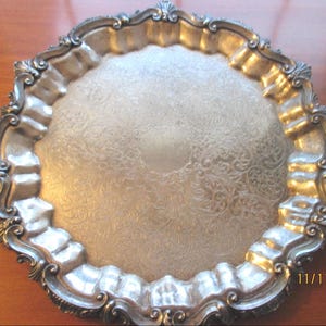 May include: Ornate silver-toned serving tray with a detailed floral pattern. The tray has a scalloped edge with decorative embellishments. A classic piece for serving or display.