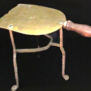 May include: Antique brass iron rest with a wooden handle. The iron rest has a curved, leaf-shaped top and is supported by three slender, curved legs. The handle is a dark brown color.