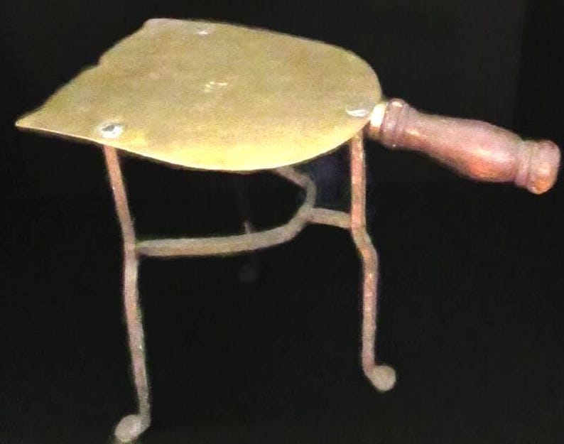 May include: Antique brass iron rest with a wooden handle. The iron rest has a curved, leaf-shaped top and is supported by three slender, curved legs. The handle is a dark brown color.