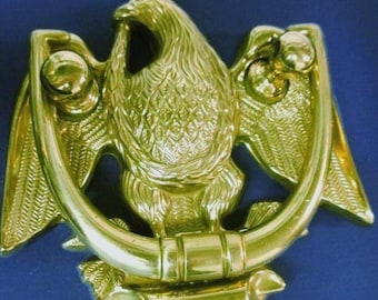 May include: A polished brass door knocker shaped like an eagle with its wings spread wide. The eagle is perched above a curved, horizontal bar that serves as the striking element. The metal surface is reflective and features detailed feather textures.