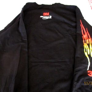 May include: Black long-sleeved shirt featuring a flame design in red, orange, and yellow on the right sleeve. The shirt displays the "3M" logo in red on the sleeve and back.