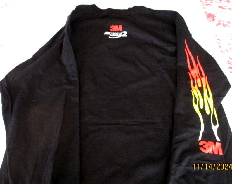 May include: Black long-sleeved shirt featuring a flame design in red, orange, and yellow on the right sleeve. The shirt displays the "3M" logo in red on the sleeve and back.